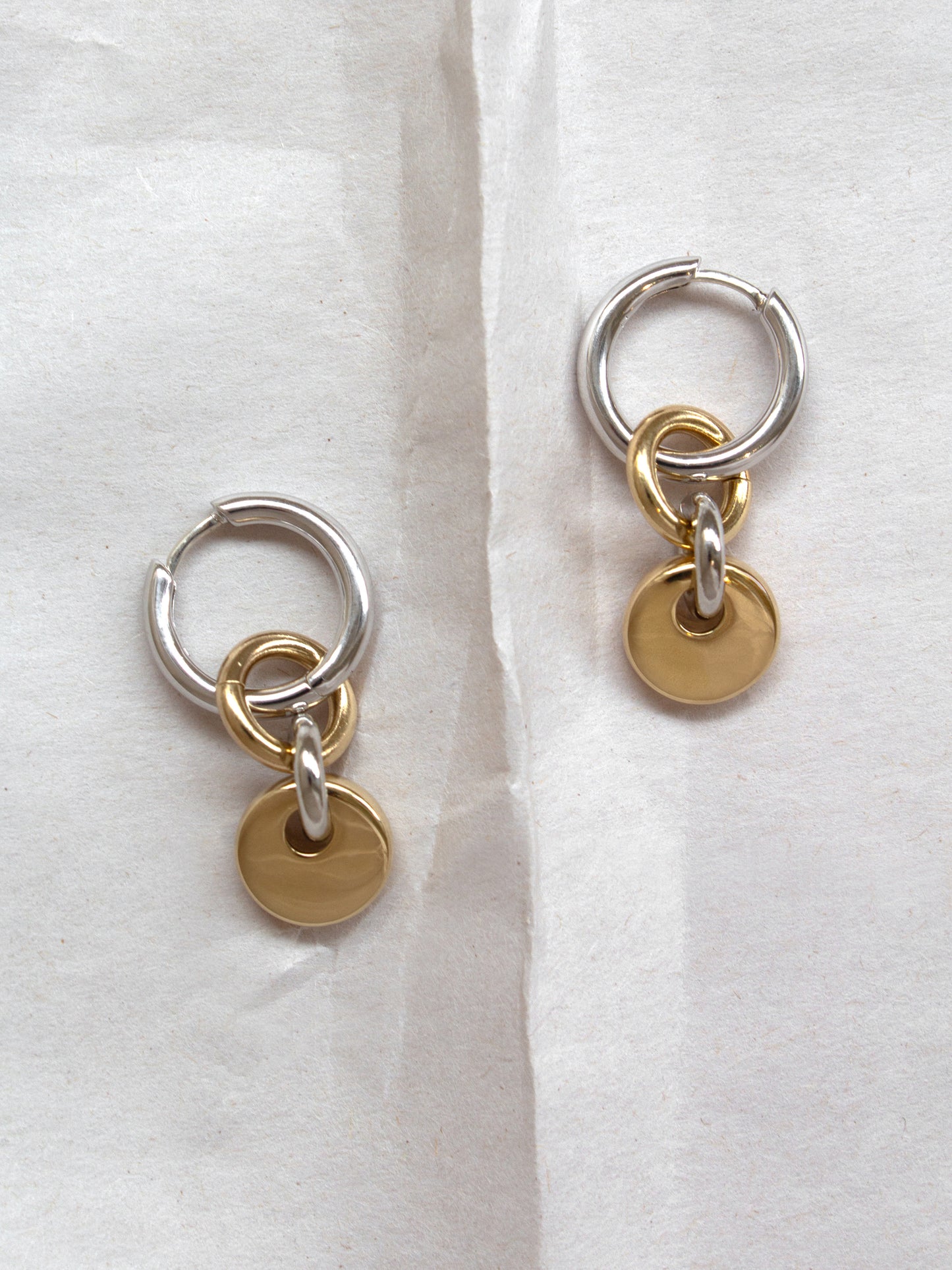 Pair of silver hoops with gold rectangular charms laid flat on a paper background.