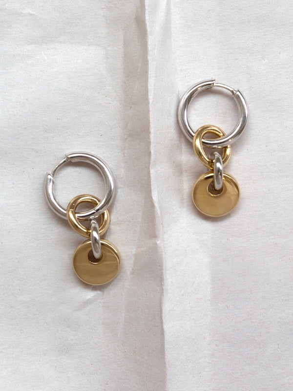 Pair of silver hoops with gold rectangular charms laid flat on a paper background.