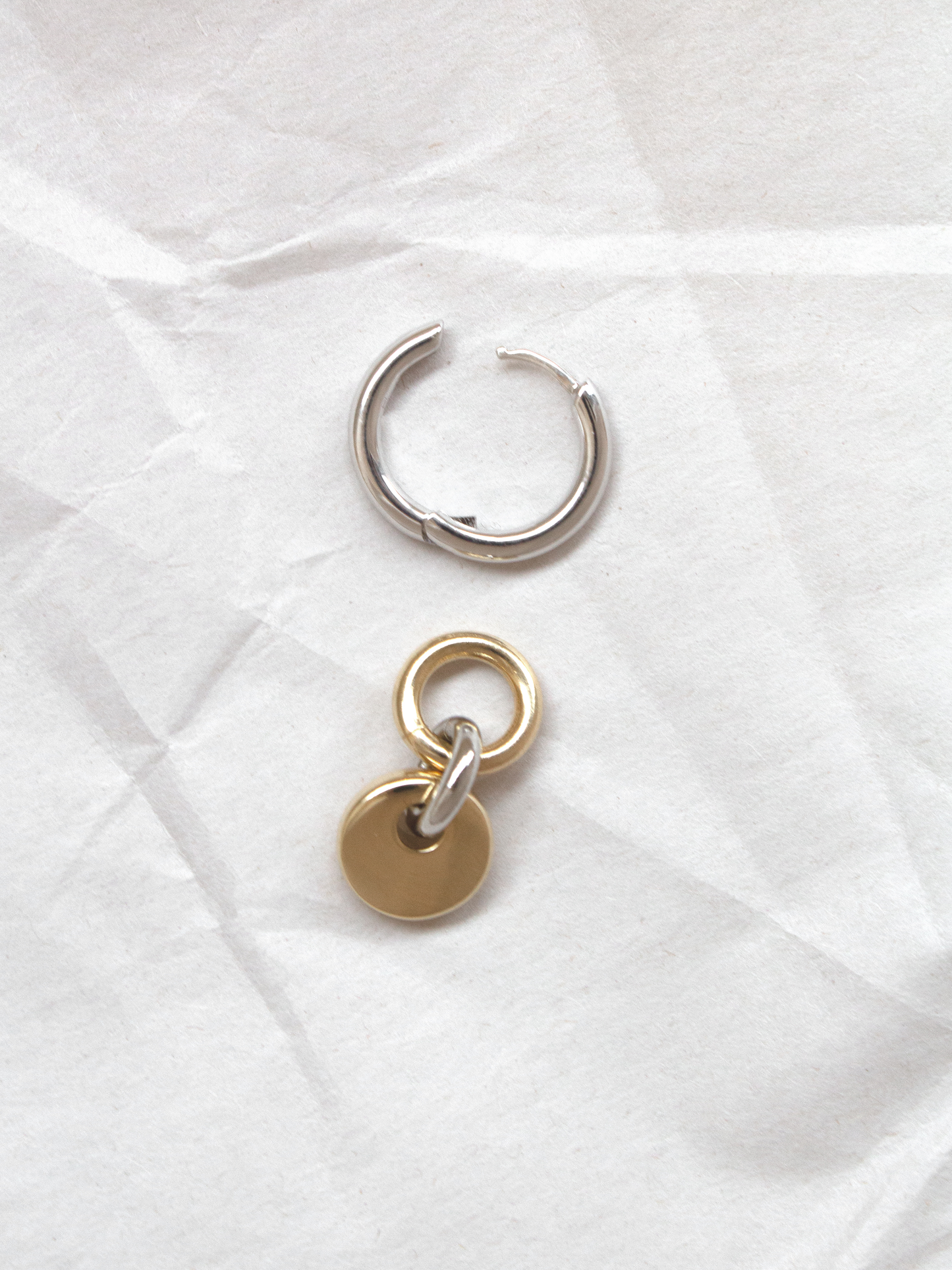 A single silver hoop with removable gold circular charm shown separately, laid flat on a paper background.