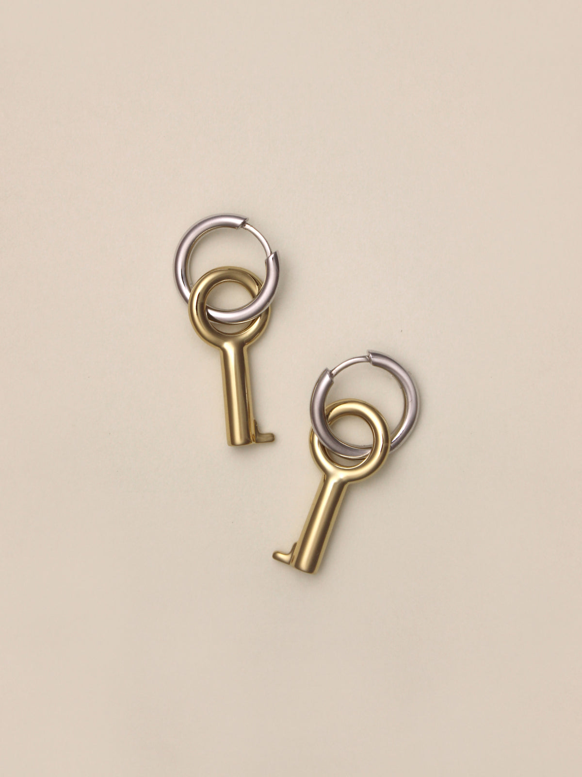 Laura Lombardi Two Tone Entra Earrings, a pair of petite silver hoop earrings with dangling gold key charms, laid askew on a neutral background.
