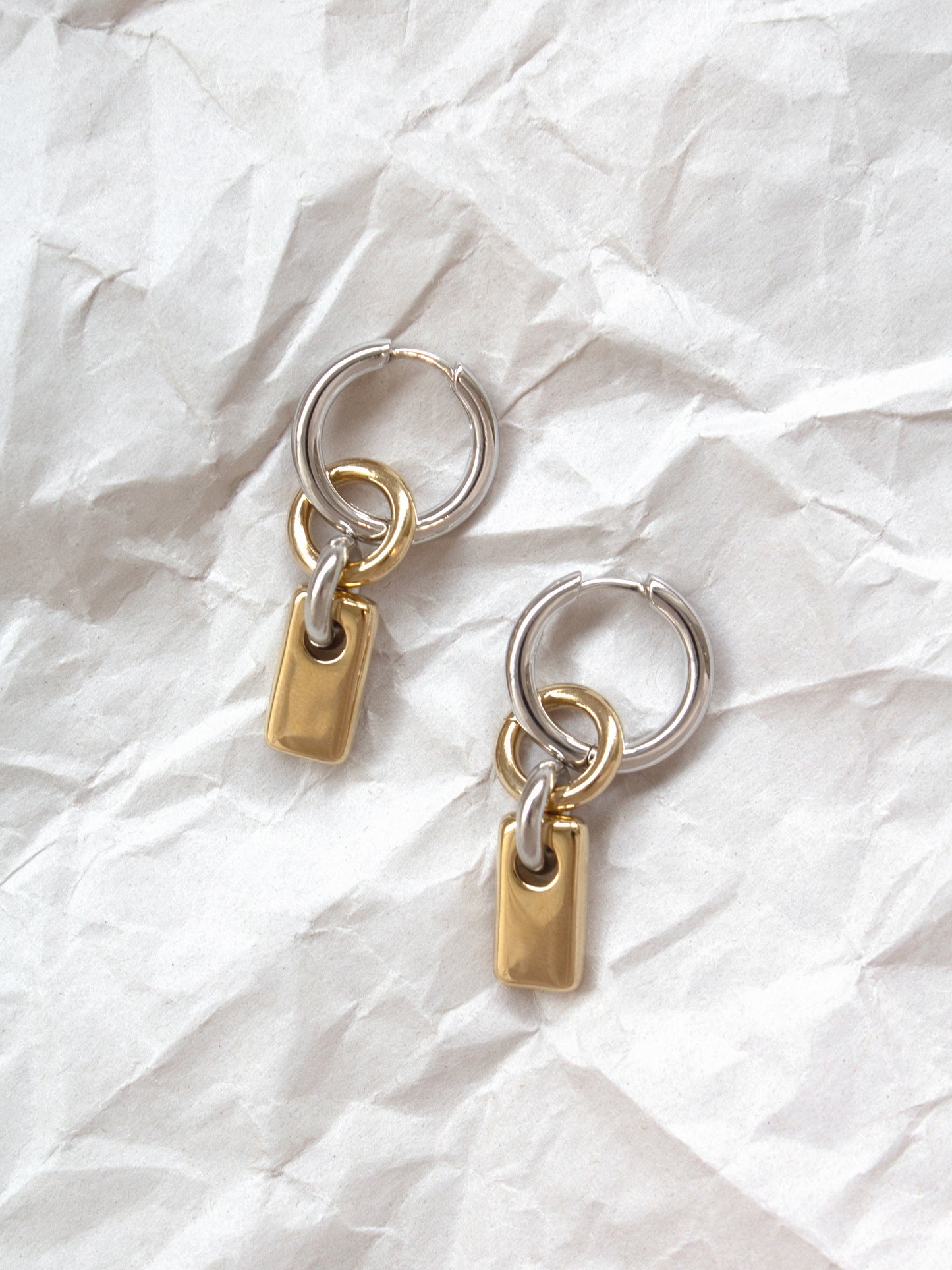Pair of silver hoops with gold rectangular charms laid flat on a paper background.