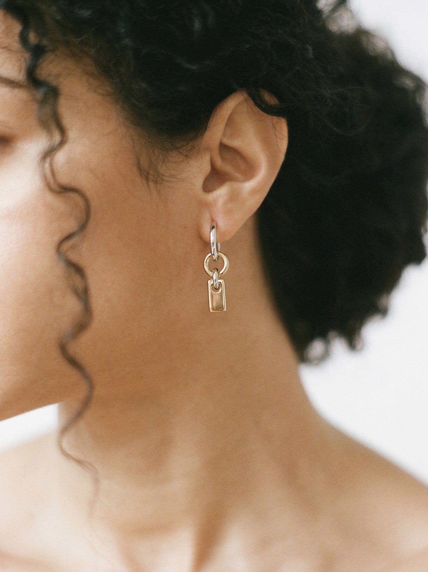 Model wearing a pair of silver hoops with gold rectangular charms on a neutral background.