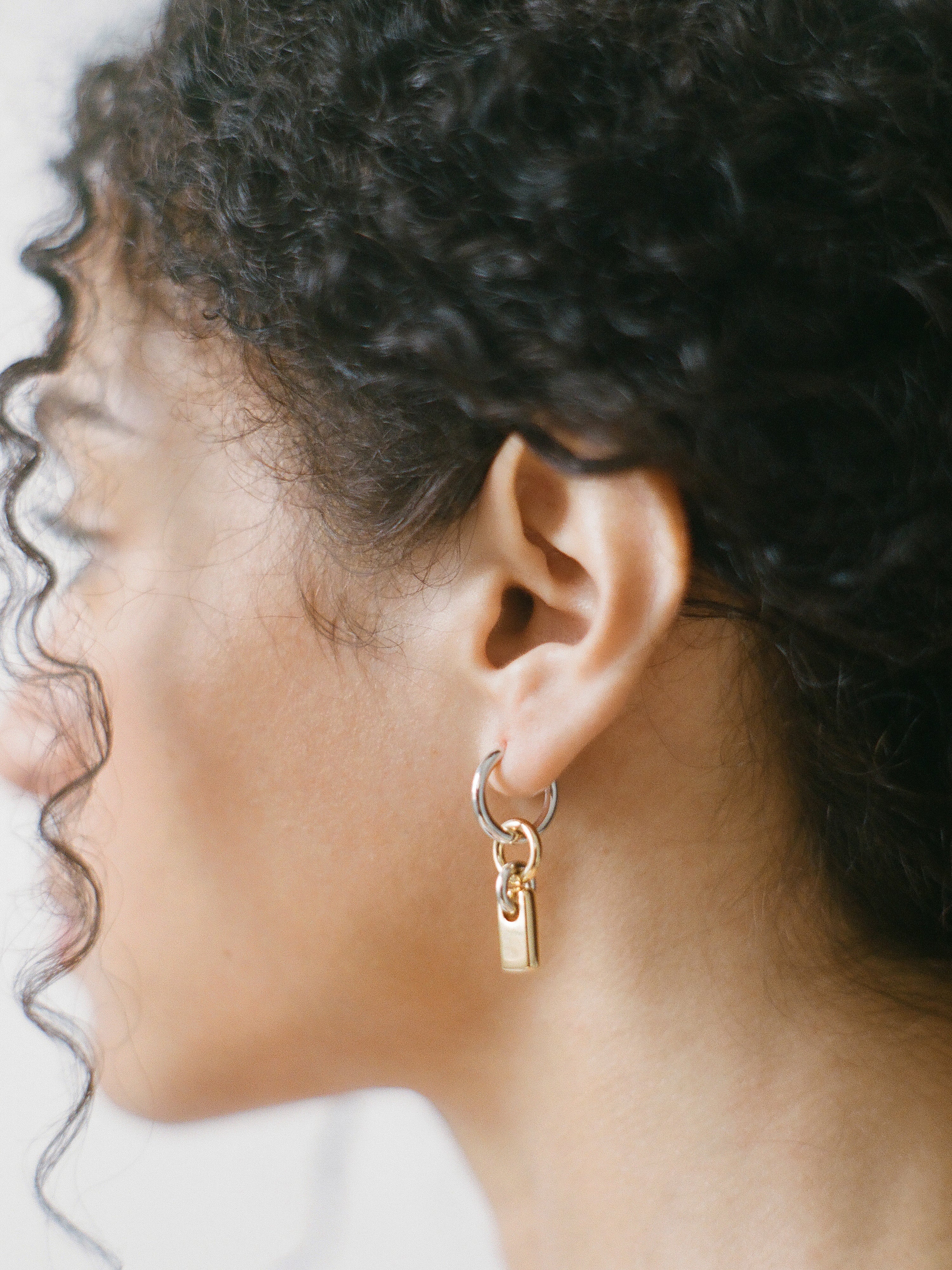 Model wearing a pair of silver hoops with gold rectangular charms on a neutral background.