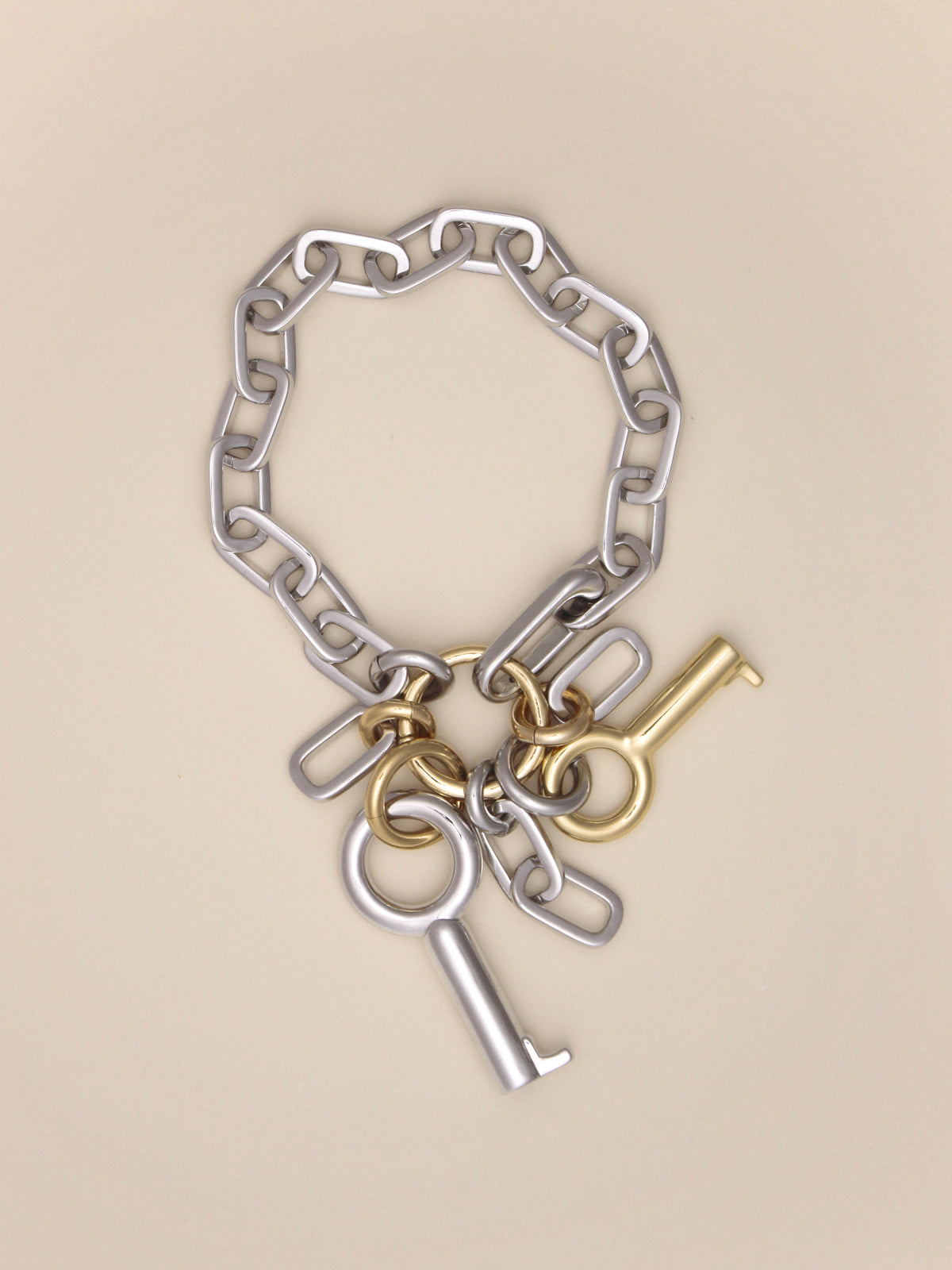 Laura Lombardi Permesso Bracelet, a silver chain bracelet with hanging keys and  chains in mixed silver and gold finishes, on a neutral tan background.