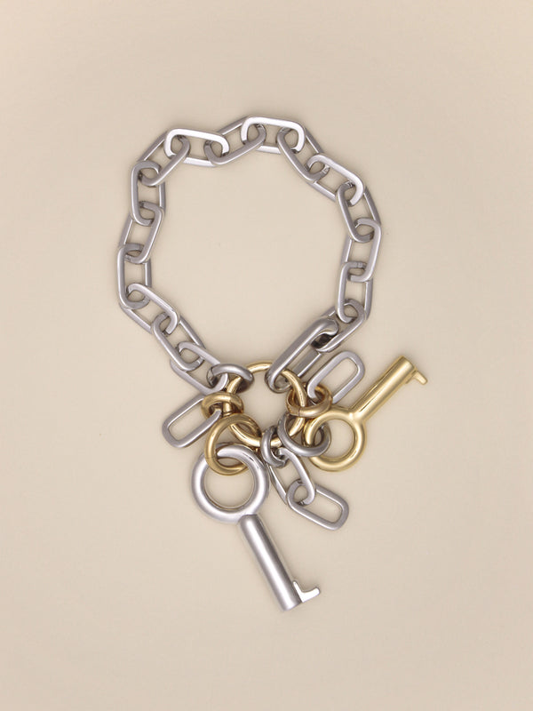 Laura Lombardi Permesso Bracelet, a silver chain bracelet with hanging keys and  chains in mixed silver and gold finishes, on a neutral tan background.
