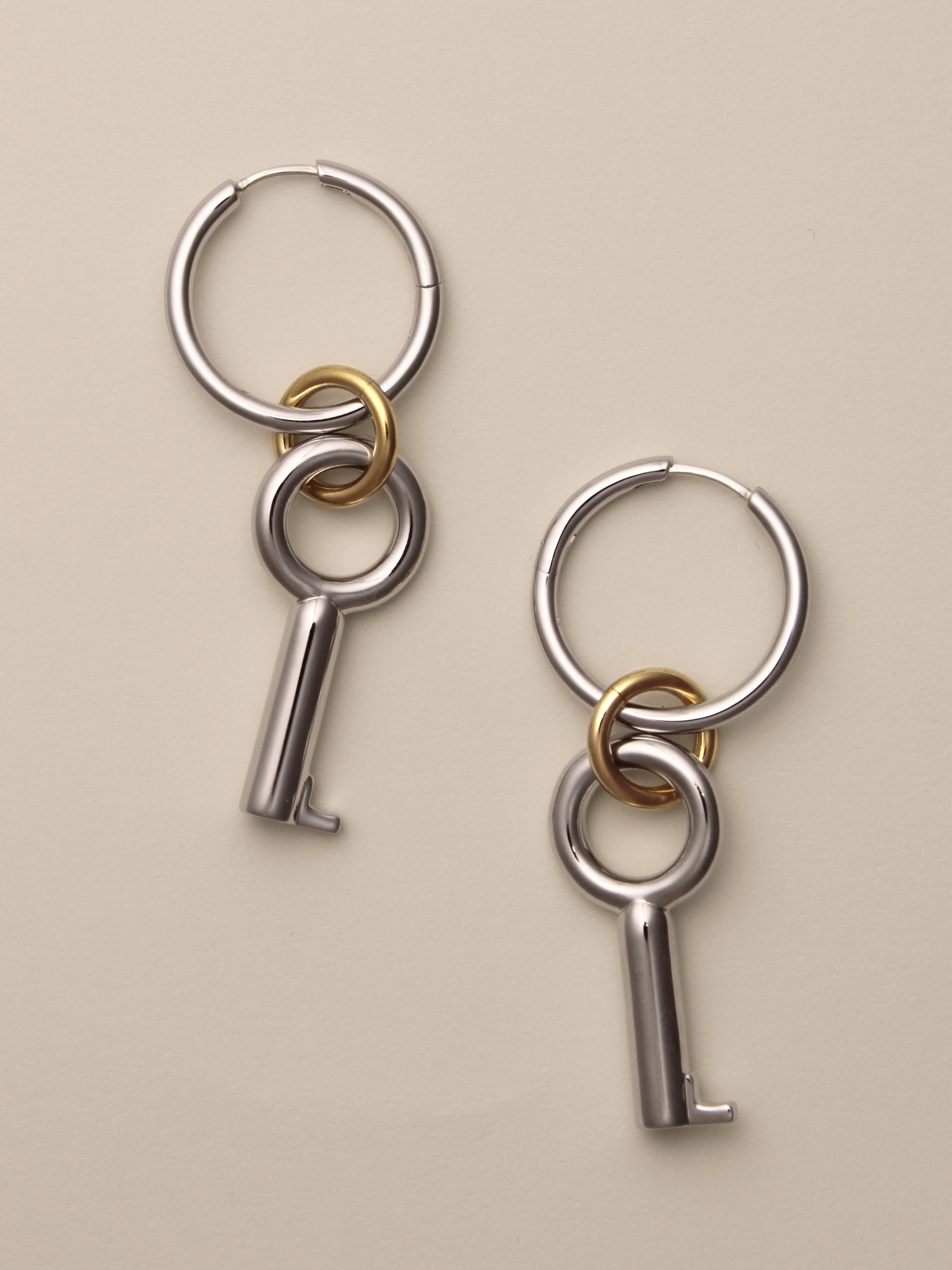 The Two Tone Permesso Earrings, a pair of silver hoop earrings with key-shaped charms and small gold rings connecting the hoops, shown on a neutral background.