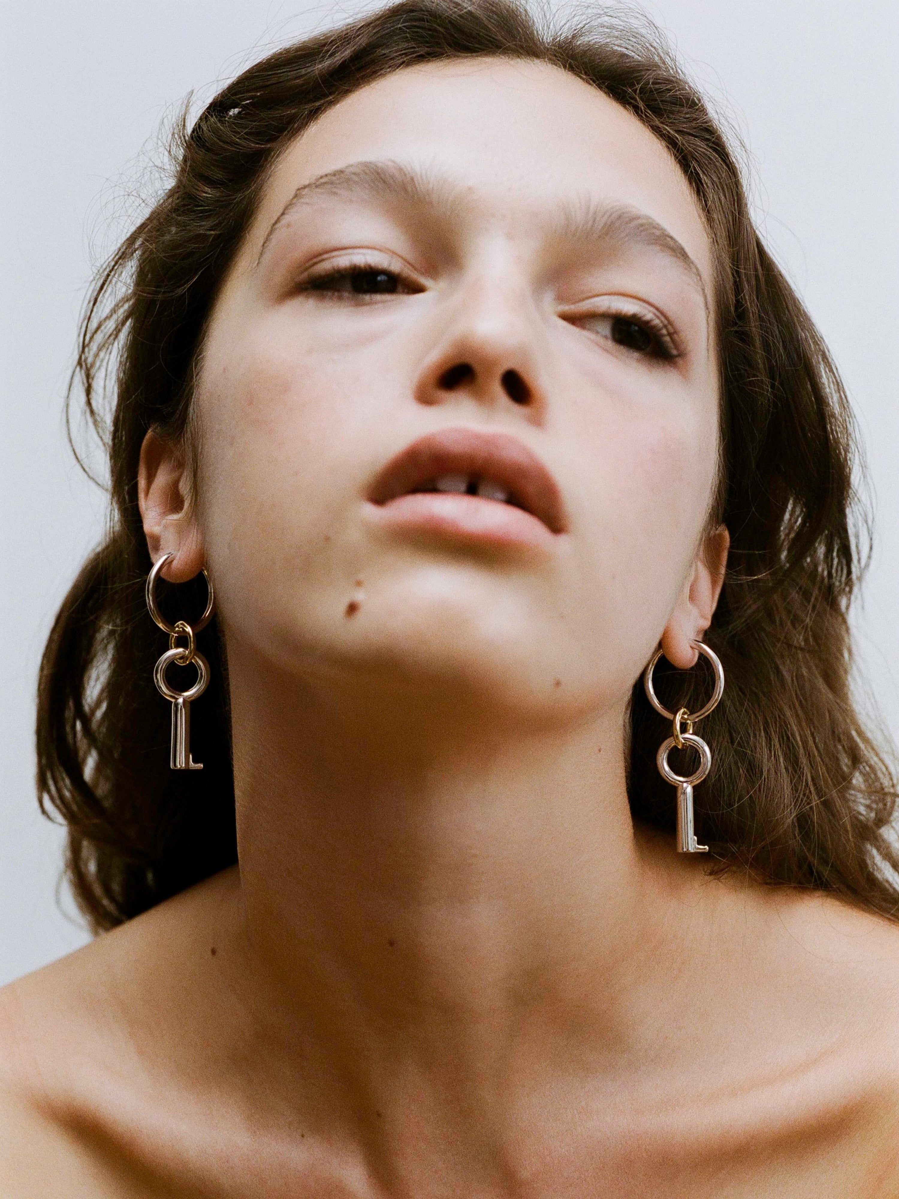 Laura Lombardi Permesso Earrings, a pair of silver hoop earrings with key-shaped charms and small gold rings connecting the hoops, shown on a plain white background.